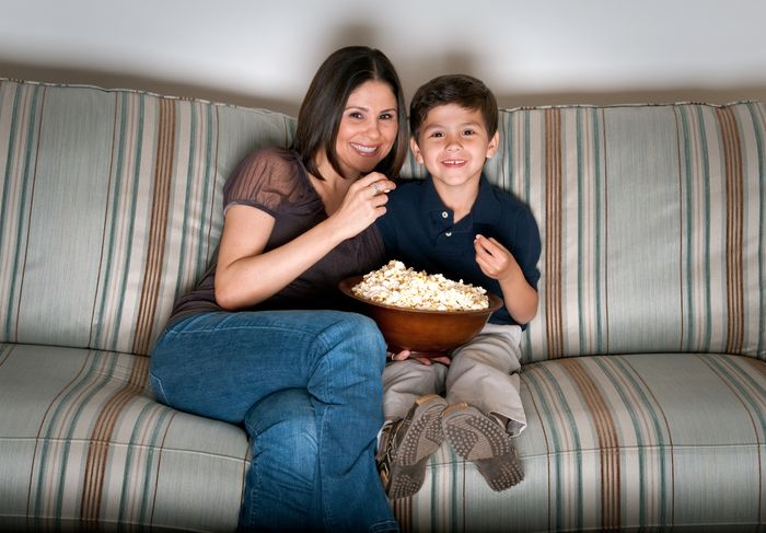 A mother and son sit on a couch, eating popcorn and watching a movie together.